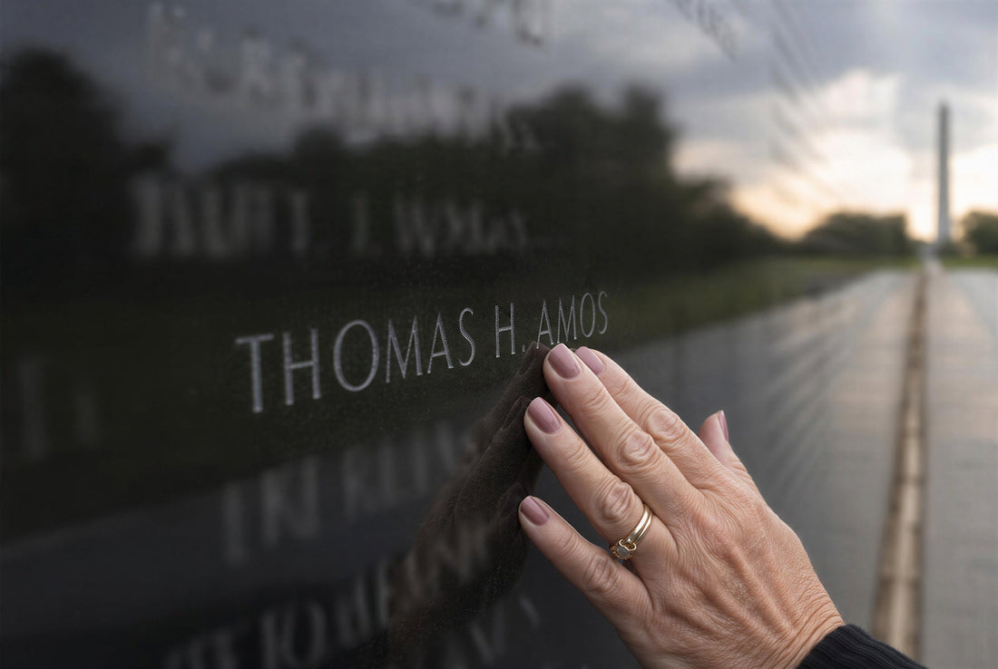 A hand touching the name Thomas Amos on the Vietnam Veterans Memorial Wall in Washington D.C., with the Washington Monument reflected in the black granite at dawn — a tribute to a fallen soldier and the welcome home he deserved.