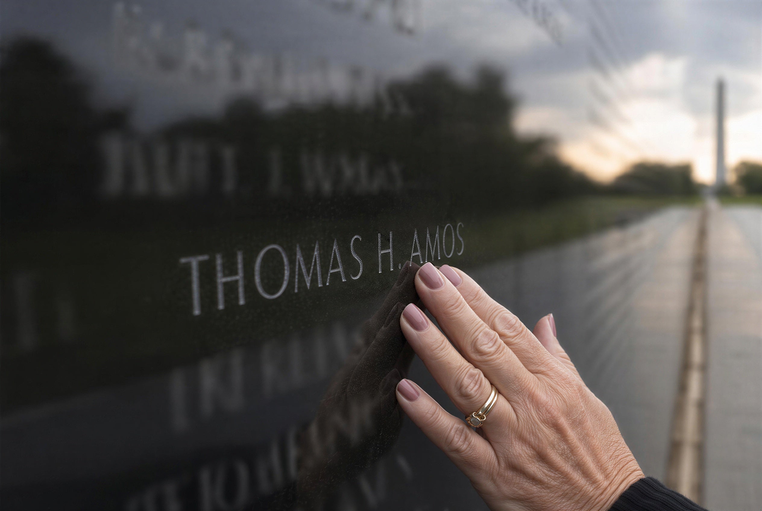 A hand touching the name Thomas Amos on the Vietnam Veterans Memorial Wall in Washington D.C., with the Washington Monument reflected in the black granite at dawn — a tribute to a fallen soldier and the welcome home he deserved.