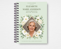 Product mockup of the Sage & Ivory memorial guest book — soft sage green cover with ivory gardenias and eucalyptus sprigs framing a gold diamond portrait frame, name and dates in dark serif type, and a sentiment line along the bottom edge.

